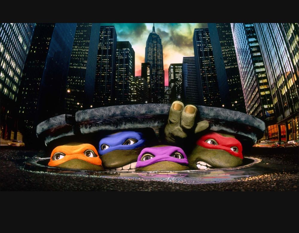 Image is of a movie poster for the Ninja Turtles movie. The backdrop is of a city. The four Ninja Turtles characters can be seen peeking out of a sewer manhole at the bottom of the image. 