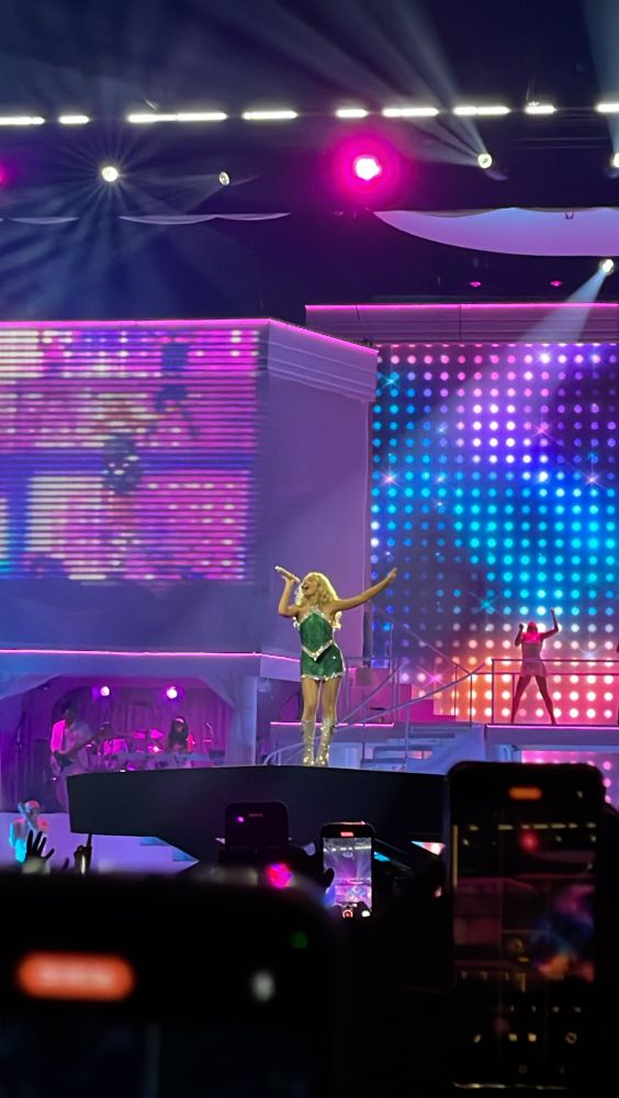 sabrina carpenter in green halter skirt and mini skirt standing on stage singing with rainbow pixel background