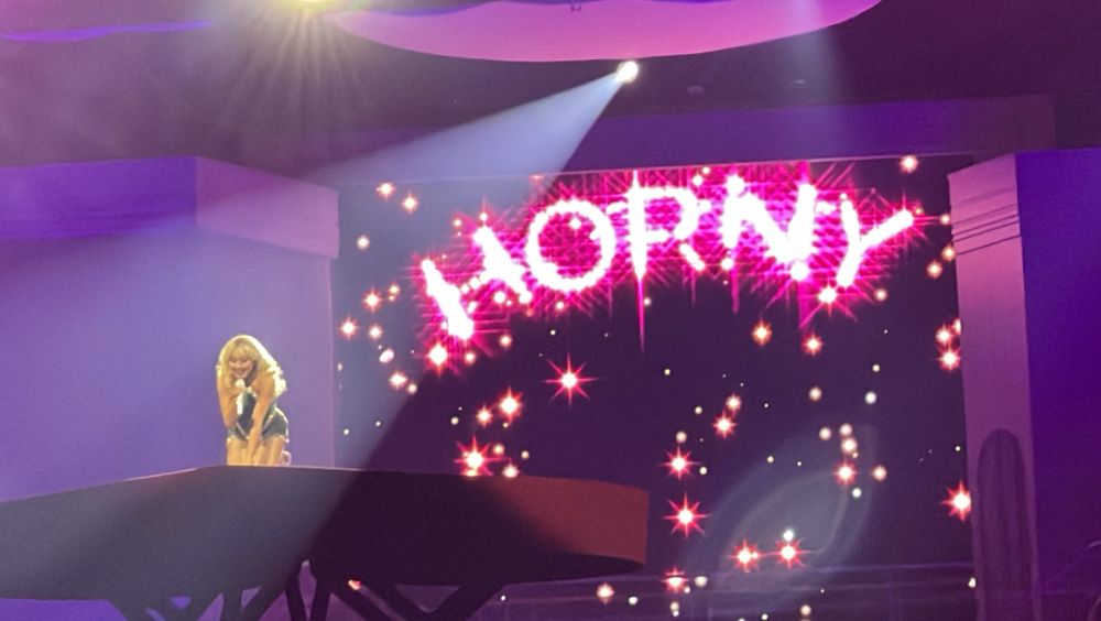 sabrina carpenter kneeling on stage with “horny” on screen behind her