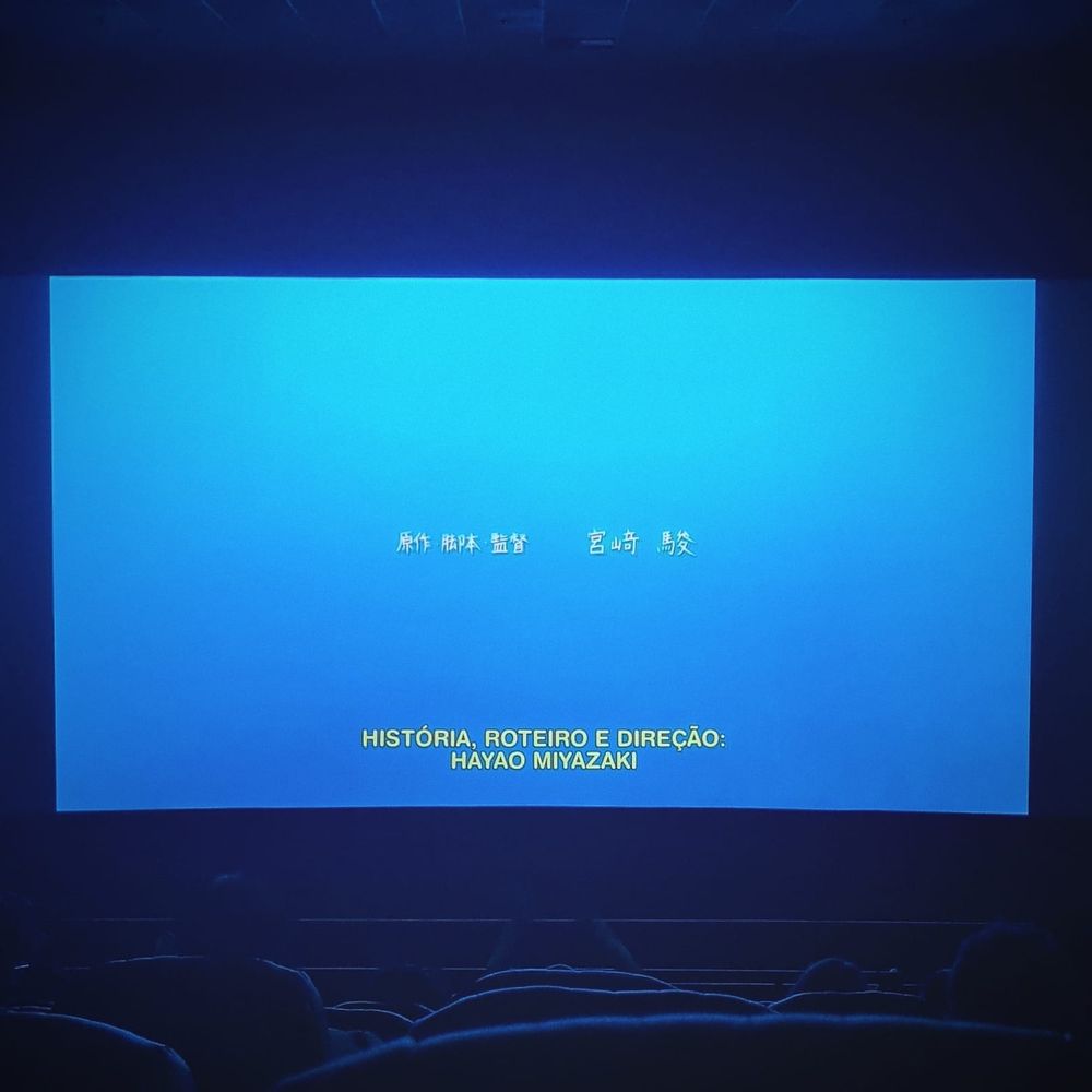 Tela de cinema com a projeção dos créditos finais do filme “O Menino e a Garça”. A tela dos créditos tem fundo azul e a legenda tem a tradução dos dizeres em japonês, creditando a história, roteiro e direção a Hayao Miyazaki.