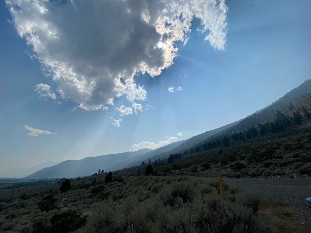The sky is blue, except there is a somewhat decent sized dark cloud hovering over the mountain slope. The sun is behind the cloud, casting a shadow on the mountain and also creating crepuscular rays. Jack's Valley in Carson Valley, Nevada.