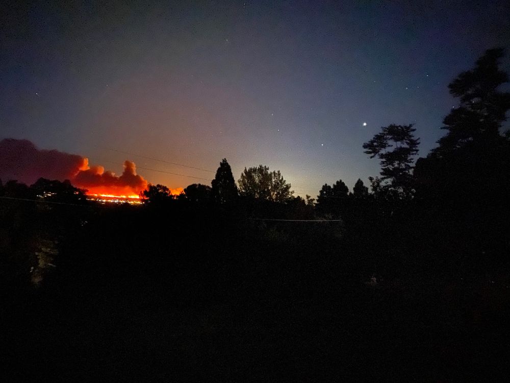 It is dark but the flames and smoke from a wildfire 15-20 miles away across the valley are lighting up the night sky. The foreground trees are silhouetted, and some stars can be seen in the sky. Carson Valley, Nevada.