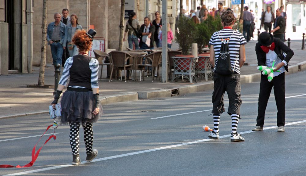 Three street performers dressed in varying black, white and red outfits are walking down the middle of a street juggling bowling pins and twirling ribbons. There is a woman with polka dot leggings and bright, naturally red hair with a small hat on top. One man is wearing matching striped socks and shirt, and the third man is in black pants, jacket and hat with a white shirt and red bow tie. People walking down the street don't seem to be paying attention, except for one man. There are cafe tables and chairs out on the broad sidewalk next to the street. Belgrade, Serbia