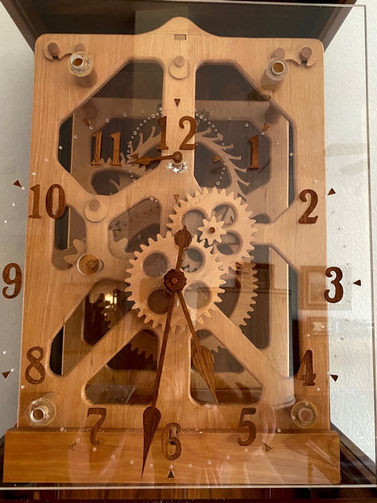 A straight-on view of the face of the wooden clock. The gears, hands, numbers, and casing are all made of wood, housed in a wood and glass case. The face of the clock is plexiglass with wooden numbers.