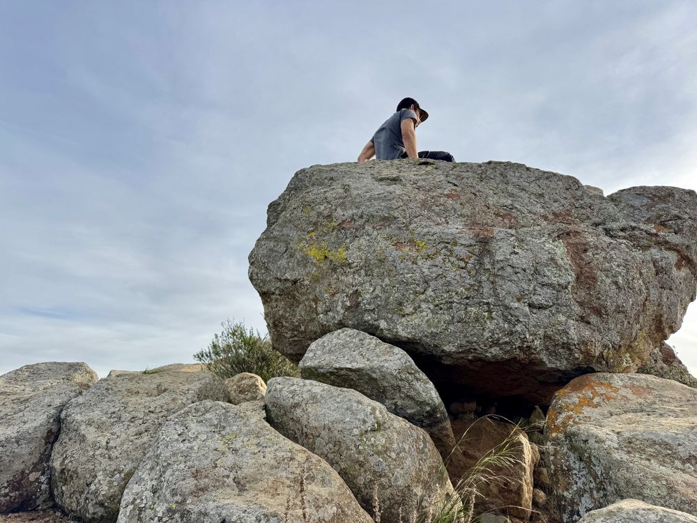 A man sits on top of one of the huge boulders on top of Cerro San Luis in San Luis Obispo, California. He has a baseball cap on and is looking down at the view below. The sky is thin clouds.