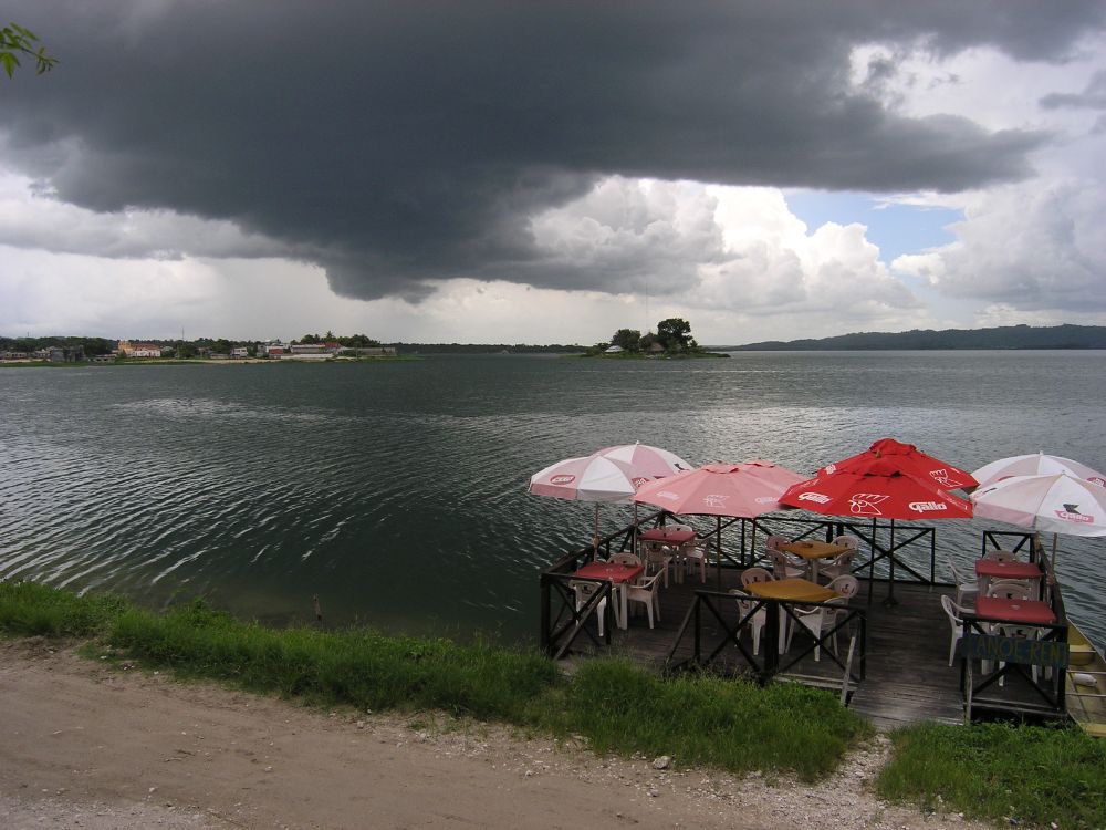 Very black clouds hang heavily over a large lake. The water looks dark as well, especially against the bright red, pink and white shade umbrellas for a small outdoor cafe on the edge of the lake.