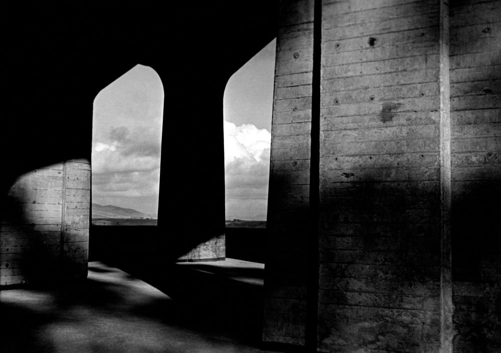 Looking through concrete pillars and arches with patches of contrasting light and shadow. The primary pillar is in the shadow and is black with puffy white clouds behind it as a strong contrast. The concrete has a wood plank texture. Geisel Library (also known as Central Library) on the University of California (UCSD) campus in La Jolla, California.