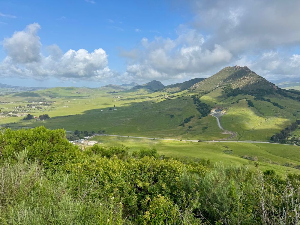 Looking at Bishop Peak from Cerro San Luis. The hills and mountains are green and there are scattered clouds in the sky, leaving patterns of light and dark.