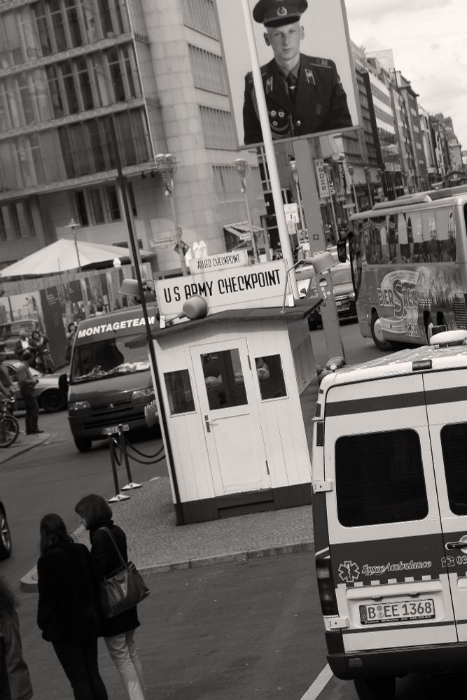 A monotone image of the well-known guard station Checkpoint Charlie which was the best-known points of passage through the Berlin Wall between the East and the West. A replica of the original guard post is in the photo (the real one is in a museum). There is traffic all around as well as people standing and walking. High rise brutalist apartment buildings are in the background. There is a photo of a soldier posted at the checkpoint. The picture of the soldier at the site of Checkpoint Charlie today is the former US army tuba player Jeff Harper. His picture is part of a series of photographs, taken to commemorate the last Allied soldiers in Berlin in 1994.