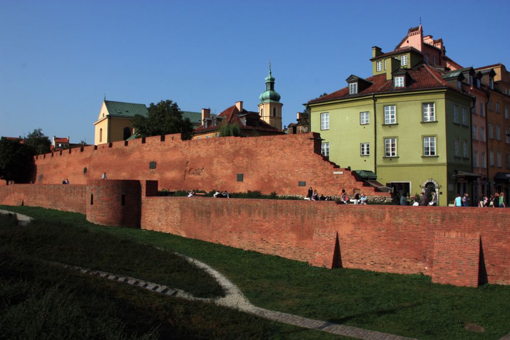 A view of the inner city of Warsaw which is built upon a massive red brick wall/fortress. One larger building in the inner city is an olive green which contrasts with red brick wall. It's later in the day so the colors are more saturated and shadows are longer. 