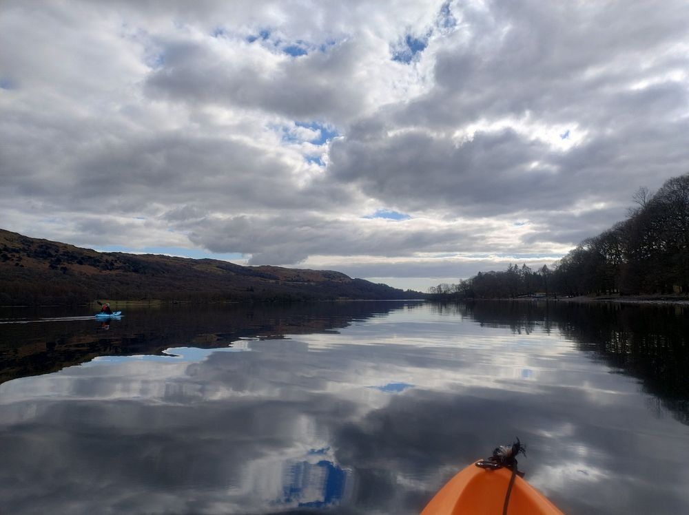 View down Coniston Water from a kayak; still water perfectly reflecting the clouds and peeps of blue sky. 
