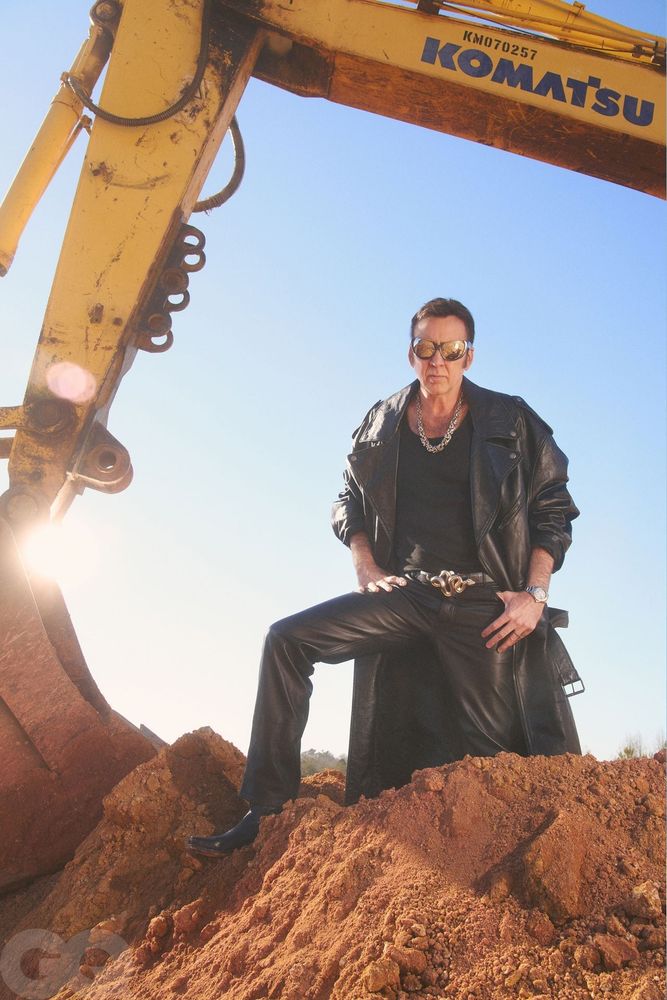 Nic Cage in sunglasses and a black leather jacket standing underneath an excavator.