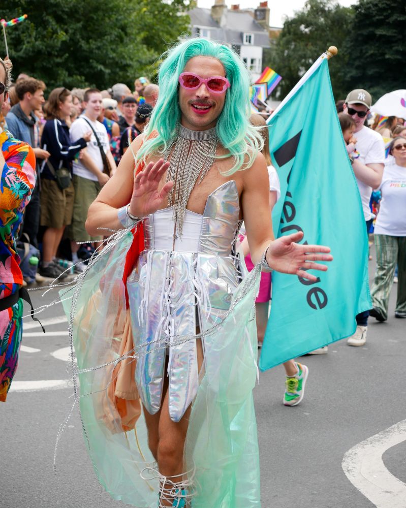 A person with cyan hair, pink shades and a silver outfit waving at the camera.