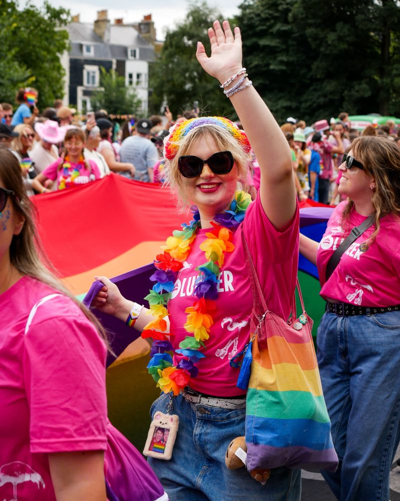 A procession holding a long rainbow banner. One of them is in a volunteer T-shirt and a rainbow lei, and is waving at the camera.