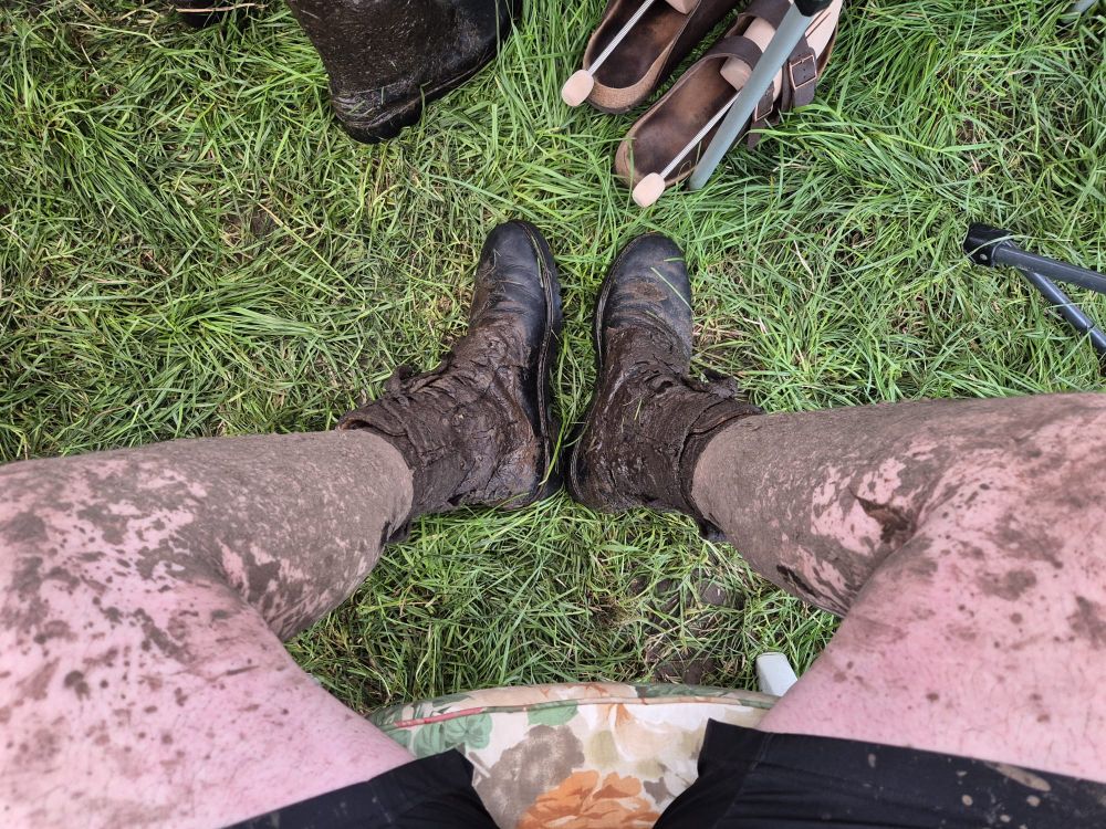 Die komplett matschigen Beine eines Wackenfans. Davor ein paar Sandalen mit Schuhspannern. 