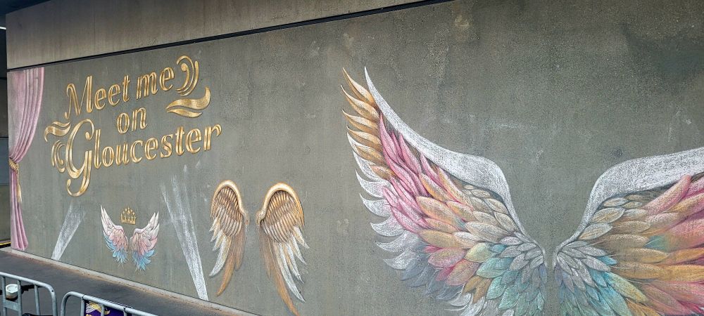 Photo is of a concrete wall with chalk street art. The words 'meet me on Gloucester' are accompanied by three sets of angel wings that people can have photos taken with.