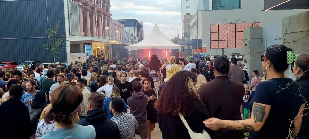 Photo is of hundreds of people dancing at a morning street party. In the distance is a gazebo with the dj and lights.