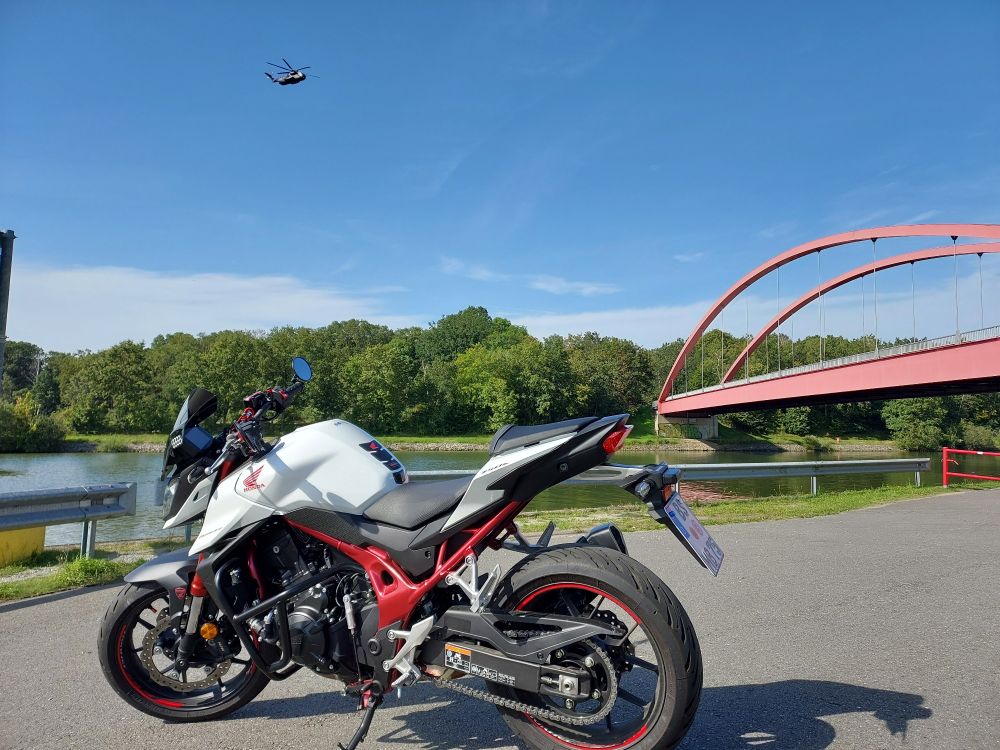 Motorrad geparkt am Kanal. Im Hintergrund eine rote Brücke, in der Luft ein Helikopter.