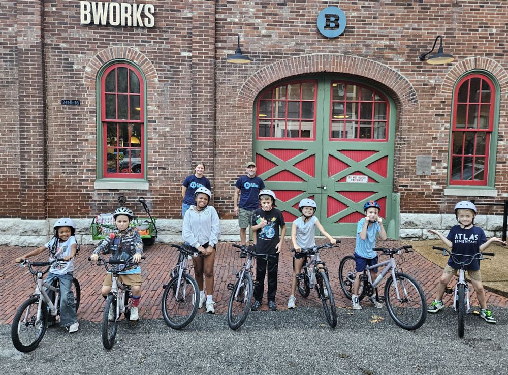 Some very proud-looking kiddos pause with their practice bikes as their dedicated BWorks teachers smile in the background after leading the group of seven Learn-To-Ride students through their pre-practice-ride safety checks.