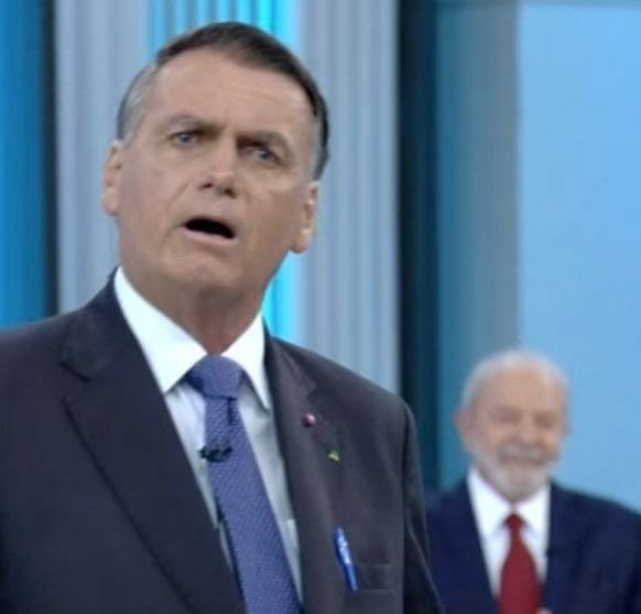 Imagem de um debate presidencial onde o Bolsonaro aparece surpreso em primeiro plano e o lula ao fundo sorrindo. Bolsonaro usa terno e uma gravata azul, Lula usa terno e uma gravata vermelha. 