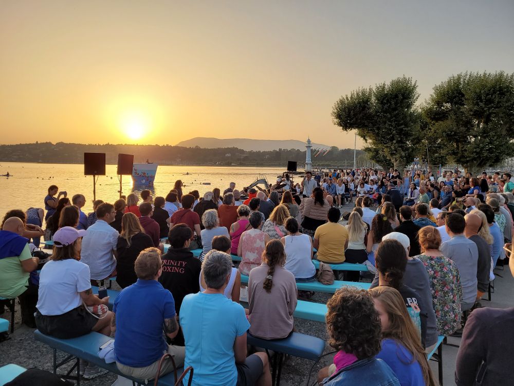 A crowd watching a band perform on the shores of a lake, with the sun rising behind