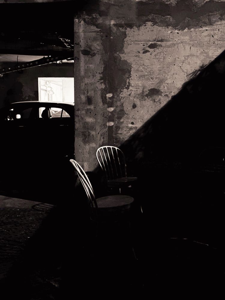 Light cast from the back lane on a concrete wall. The back s of wo patio chairs in front and the silhouette of a car behind.