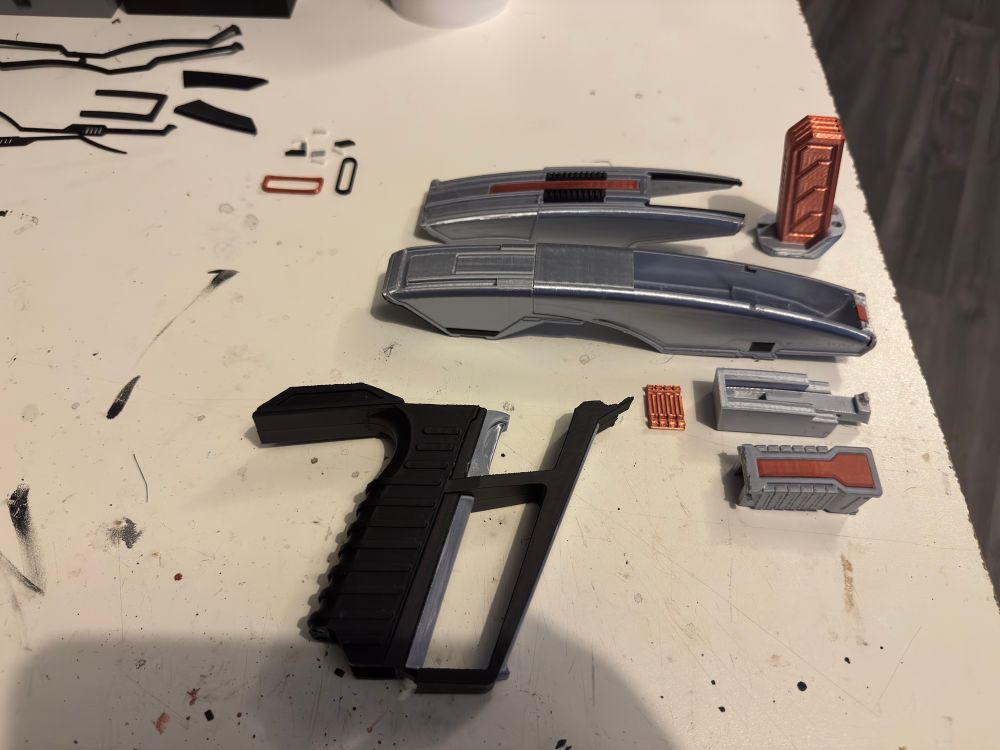 A collection of model parts displayed on a work surface, including gray and black components resembling a futuristic weapon, alongside various small detailed pieces and some copper-colored elements.