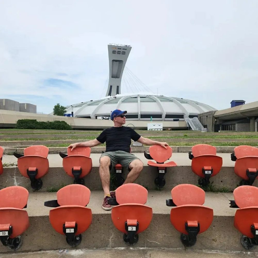 Me in shorts, t-shirt and baseball cap sitting on a red folding set in front of Montreal's Olympic Stadium.