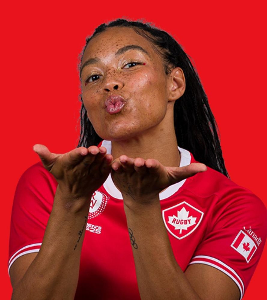 The official Rugby World Cup portrait of Canadian wing Asia Hogan-Rochester, a young woman with long black hair, brown skin and freckles. She is blowing a kiss towards the viewer and making direct eye contact.