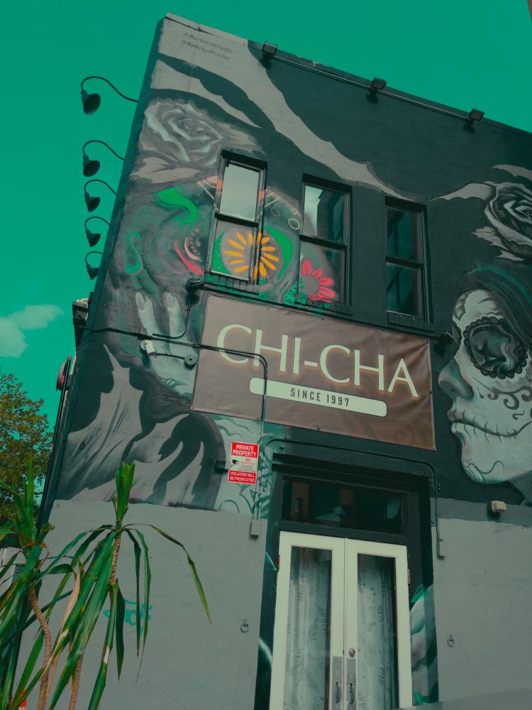 seriously cool photo of the Chi Cha lounge (since 1997). Building on an angle with sign in the center surrounded by a mural with a Mexican day of the dead style kill on one side and flowers on the other. 

Sky appears an unnatural turquoise because of the photo setting I used. They don’t call them filters. They are apparently fancier — LUT — look up table.