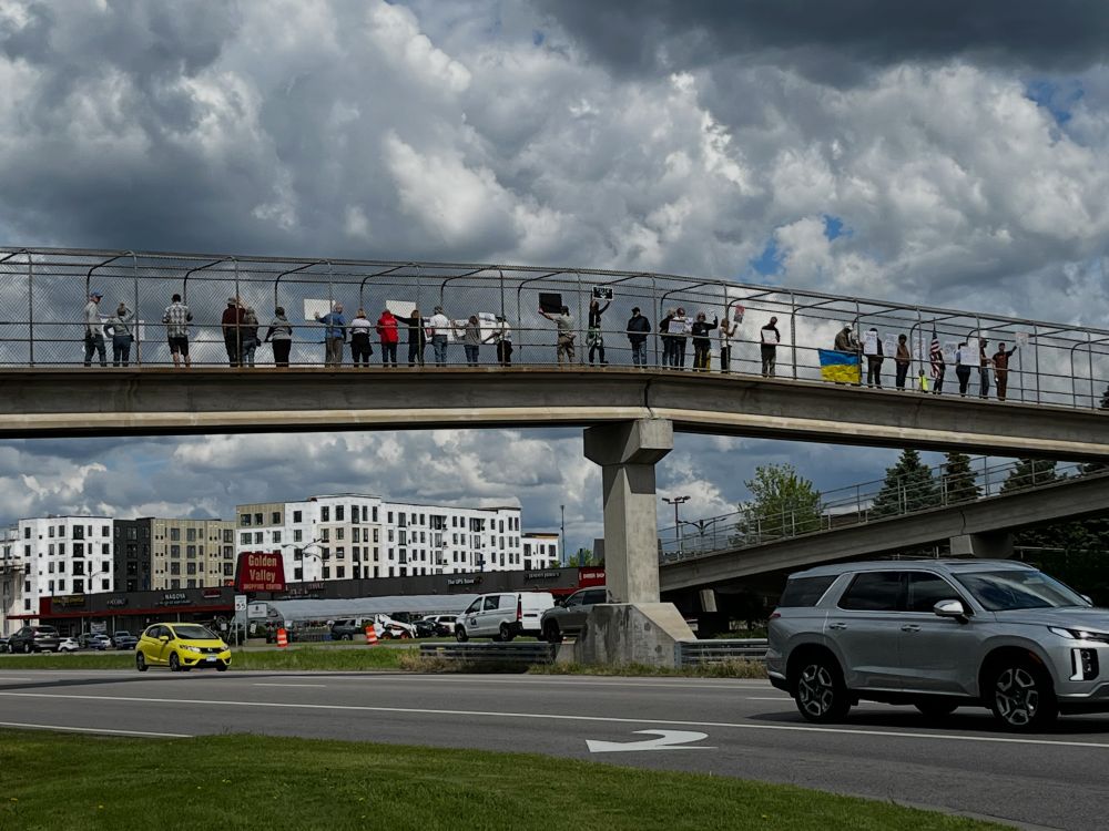 A crowd of protestors with signs on the pedestrian overpass at Hwy 55.