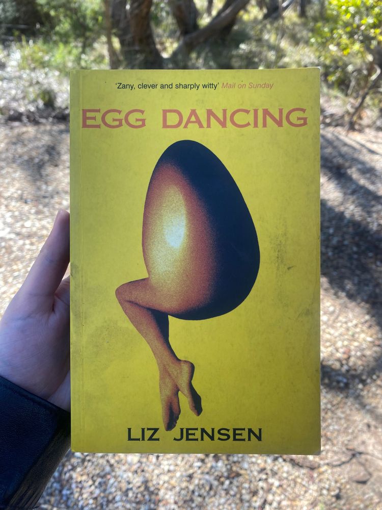 A hand holding Liz Jensen’s book ‘Egg Dancing’, the cover of which depicts an egg shell connected to a woman’s legs as one combined organism.