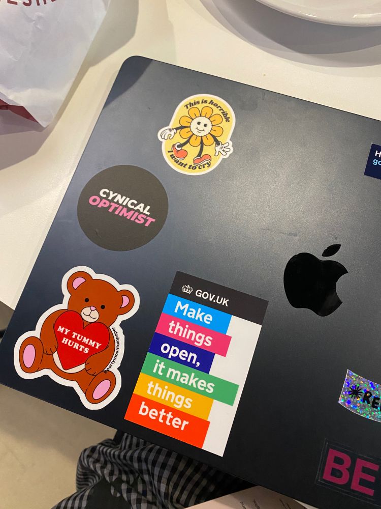 a macbook laptop decorated with a few stickers. Some stickers to highlight are a circular sticker with a black background that reads 'Cynical Optimist' with 'cynical' in white and 'optimist' in pink. Another square sticker that says 'GOV.UK Make things open, it makes things better' in vertical.