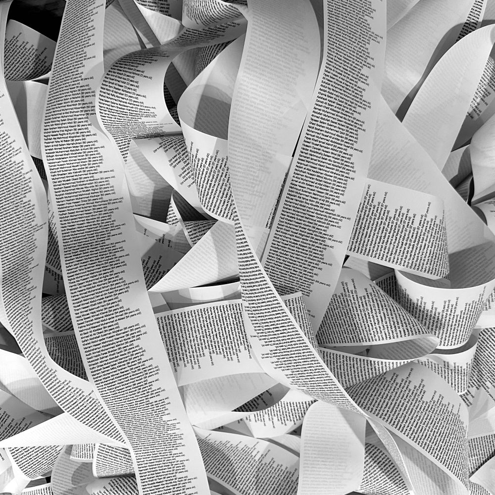 A photograph of a very long printed list of names and ages on receipt paper, piled up and curling around. Black and white. 