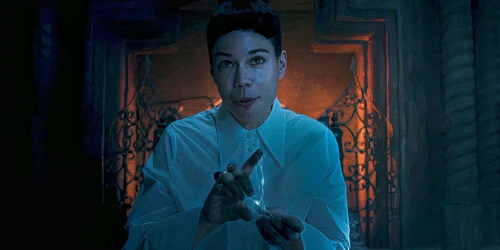 An image of Laia Costa as Moghedien in The Wheel of Time. She is wearing a white dress shirt and has her black hair in a small bun with blunt short bangs. She is sitting in front of a fireplace using small spider-web like weaves of the One Power in her hands to do magic things.