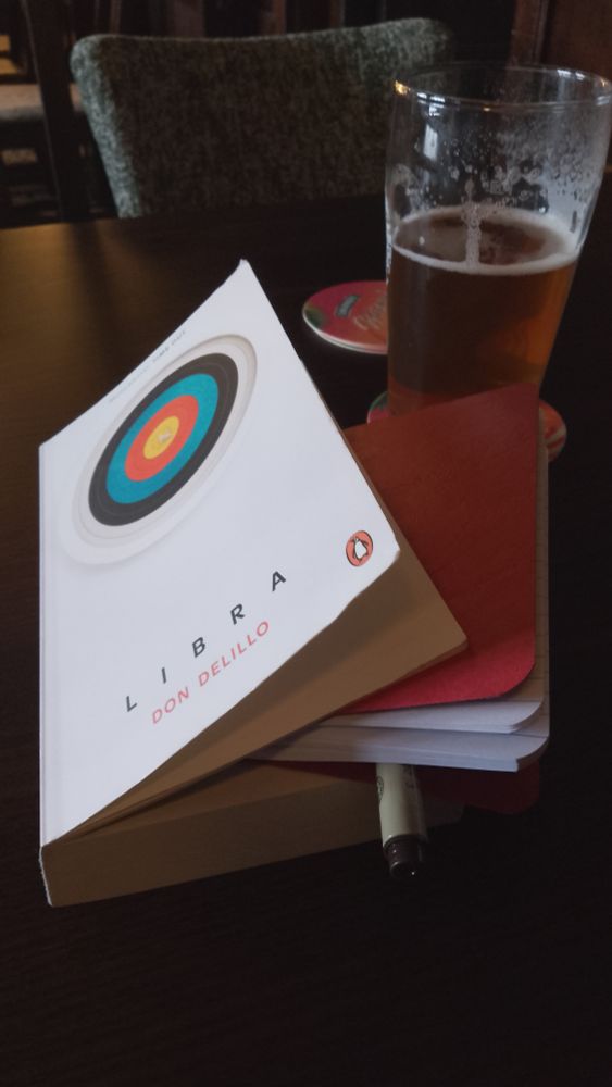 Pub table with a pint of beer, Don Delillo's book Libra and a notebook and pen being used as a bookmark