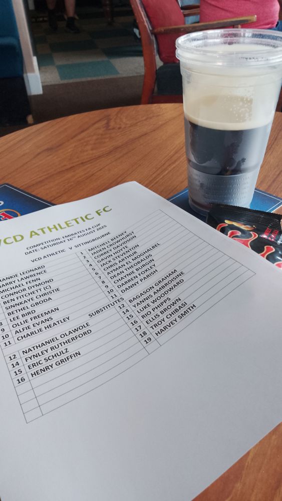 Football team sheet for VCD Athletic vs Sittingbourne, with empty peanut packet and a pint of Guinness in a plastic glass, all on a small wooden table.