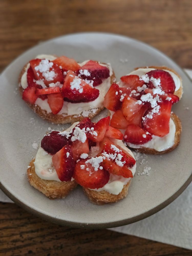 3 golden heart shaped palmier cookies topped with a smooth white cheese with bright red cut strawberries that have a dusting of white confectioners sugar on a vintage stoneware plate