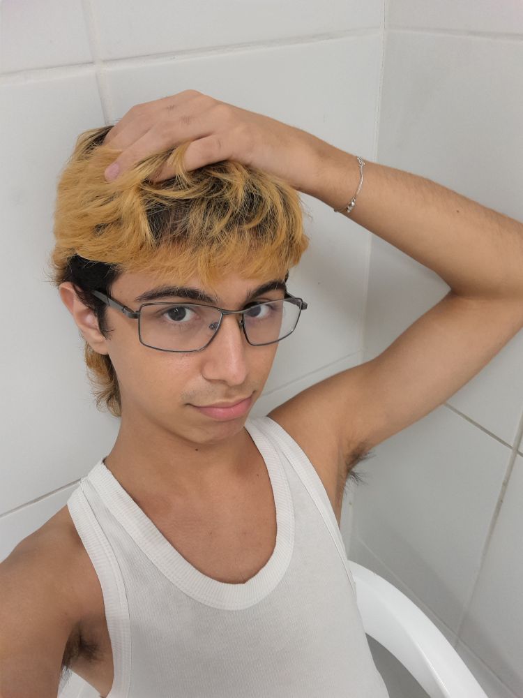 selfie de um gay magro jovem usando uma regata branca. ele tem cabelo loiro descolorido com a raiz bem crescida e uma pulseira fina prateada