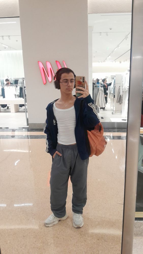 selfie de espelho de mim, andre, usando um tenis branco tratorado, uma calça de tactel cinza e uma camisa branca meio transparente e apertada. em cima disso, uma jaqueta azul escuro da adidas e uma bolsa cor pessego. meu cabelo eh recem raspado e escuro como a noite. to de fone