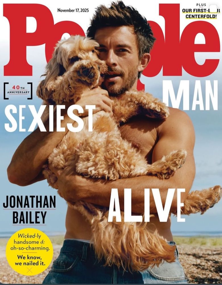 People magazine cover of Jonathan Bailey as the sexiest man alive. He’s shirtless on the beach holding a small brown doodle mix dog in his arms. 