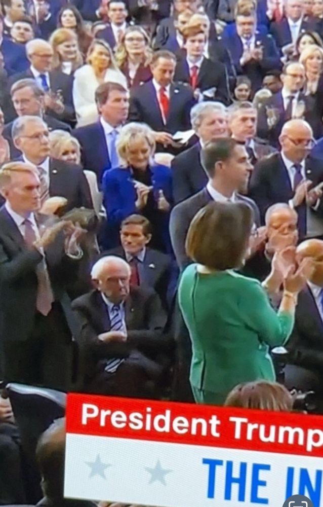 A photo from this stupid ass inauguration today, where everyone in the room is standing and applauding, but Bernie Sanders is seated center house right straight mean-mugging this fucking farce. He has his arms crossed like a malcontent and that frown can be seen from space.  He wasn’t there to support SHIT. 