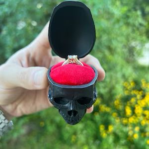 A hand holds a ring box above some grass. its a black skull that opens at the cranium to reveal a diamond engagement ring, seated in a mound of red velvet 
