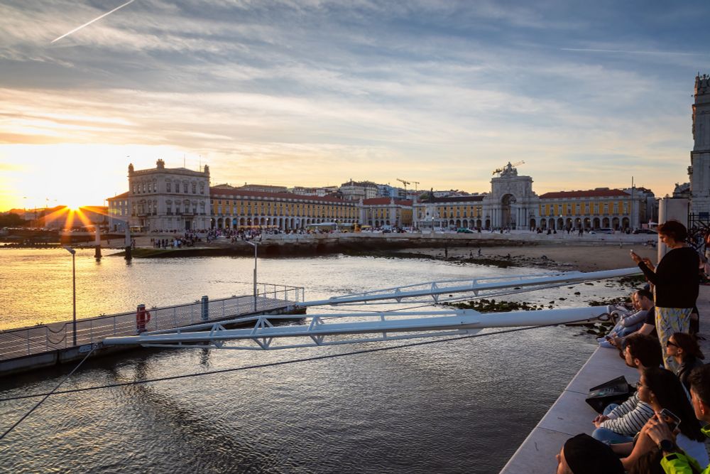 To the right are several people, most sitting, watching the sunset which is to the left. They are at water's edge. In the background, the water leads to a large plaza surrounded by elegant yellow and white buildings with columns and arches.