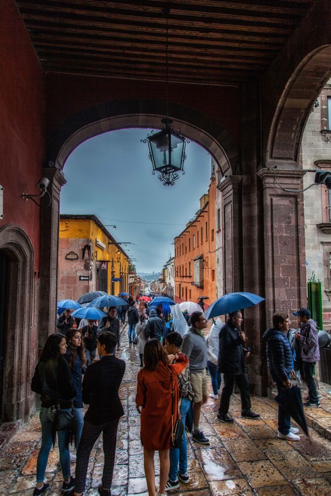 In an old Mexican city, a covered walkway with large arches has several people walking and standing. It is raining, and the paving stones are wet. Many people are waking in the street holding umbrellas, the great majority of which are blue. 