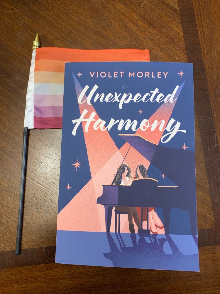 Sapphic book Unexpected Harmony which is of two woman playing a piano. Blue cover with pink spot light. A sapphic flag behind the book 