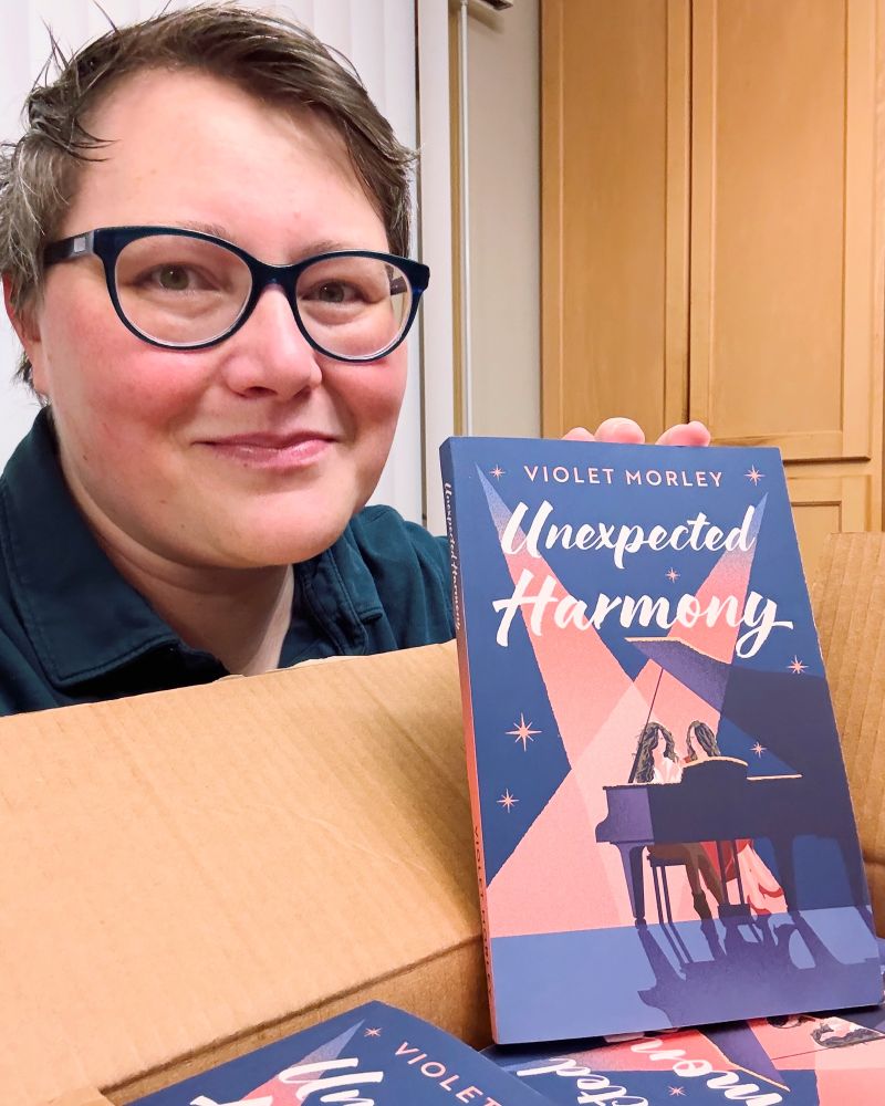Shy author holding up sapphic book called Unexpected Harmony. 
