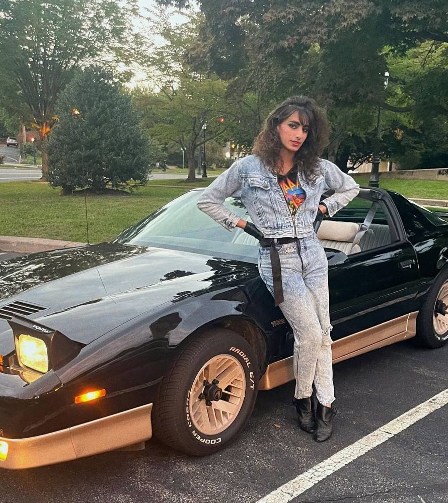 [Image ID: A woman with dark brown perm'd hair stands and leans against a 1987 Pontiac Trans AM. Her hands are one her hips as she gives a slight smile yet neutral expression to the camera. She is wearing an all denim outfit, both pants and jacket that is slightly closed. With black gloves, shoes and belt. She is wearing a black shirt that has some oranges and blues on it, but indescribable what exactly the image is. End ID]