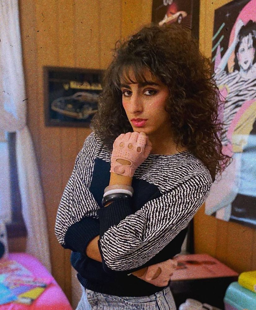 [Image ID: A woman with dark brown perm'd hair stands and leans slightly back with one arm around her mid drift while the other is bent upwards towards her face with her chin resting against the back of her knuckles. She is in an 80's themed bedroom, vibrant posters of glam rock bands and poster of cars from that area are scattered across its wood panel walls. To the right of her there are magazines and a boombox in the background, resting on a table near a window with white drapes. Along with a blue and teal makeup kit, a white hair dryer and another magazine to the left of her. The woman is wearing denim jeans, and a sweater with solid black on the abdomen but the arms and upper chest are zebra striped. She has on pink finger less gloves and three bracelets; black, white and cream. End ID]