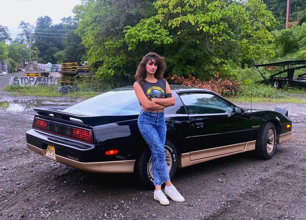 [Image ID: A woman with dark brown perm'd hair stands and leans against a 1987 Pontiac Trans AM. Her hands are crossed she gives a slight smile yet neutral expression to the camera. She is wearing black gloves, dark blue denim jeans, a sleeveless crop-top of the guns & roses band logo, along with white shoes and socks. End ID]